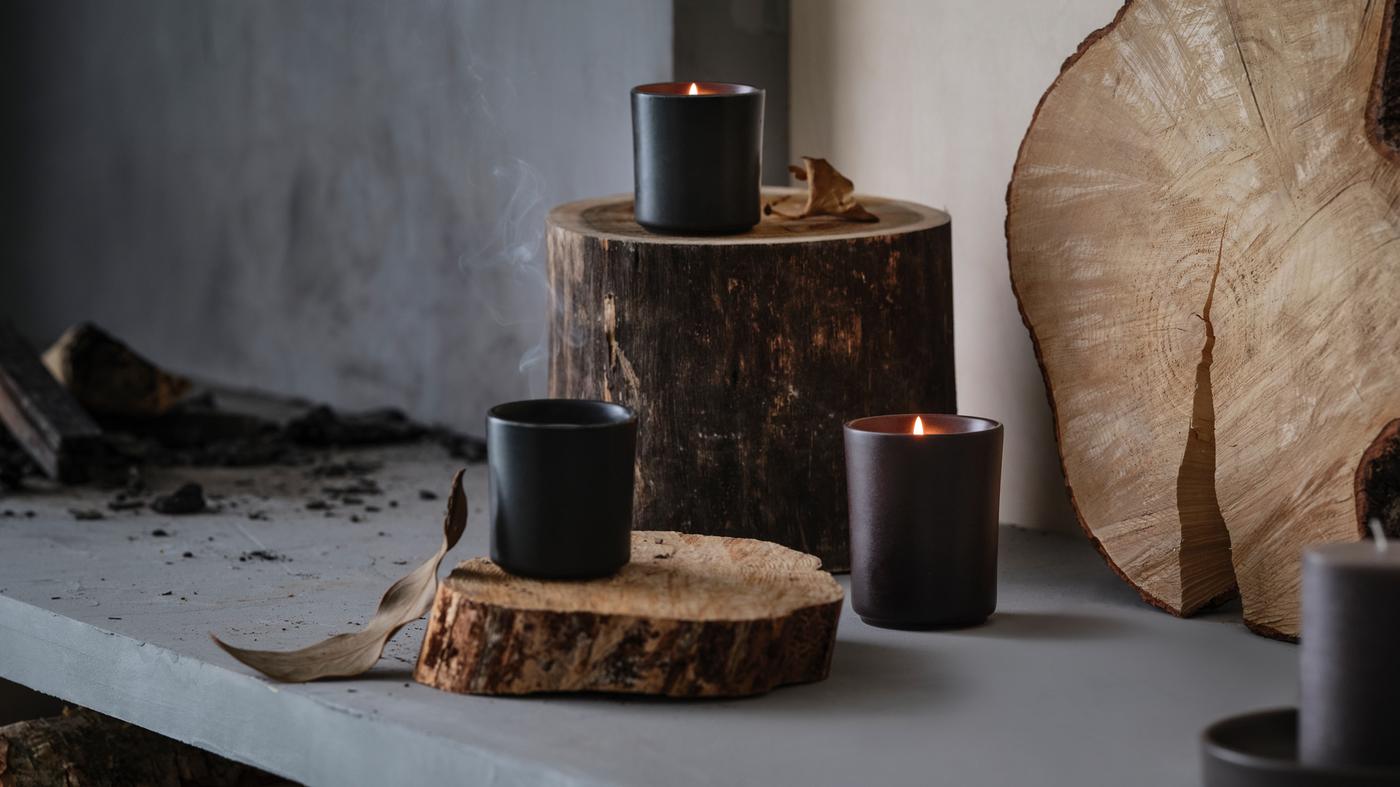 Tres candeas aromáticas BEHJÄRTAD en candeeiros de cerámica colocadas sobre uns troncos nunha cheminea de pedra rústica con anacos de madeira.