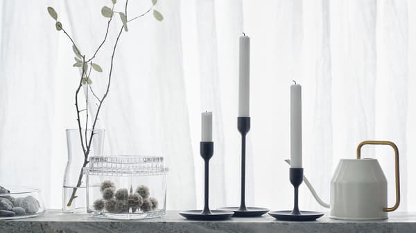 Tres candelabros negros de distintas alturas sobre un fondo branco cunha regadeira e tarros de vidro.