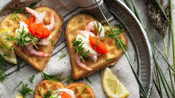 Tres gofres VÅFFLOR en forma de corazón con gambas, maionesa, perlas de algas e aneto nun prato cun pano de cociña.