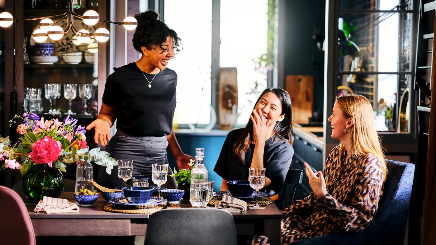 Tres mujeres riendo alrededor de una mesa de comedor con tazones ENTUSIASM, copas de vino SÄLLSKAPLIG y una jarra.