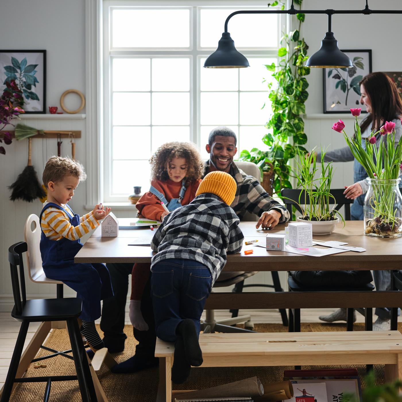 Tres niños pequeños y sus padres se mueven alegremente en una zona de comedor con una mesa KLIMPFJÄLL de color gris-café.