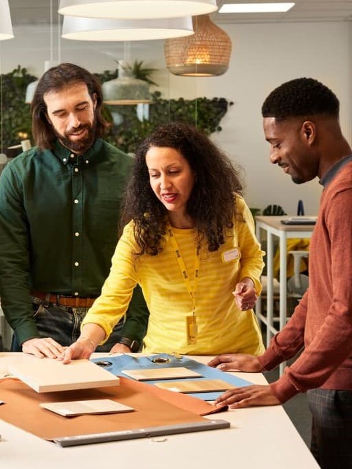 Tres persones treballant en equip en un espai creatiu, revisant mostres de materials sobre una taula. L’ambient és càlid i professional, amb decoració natural i llums penjant.