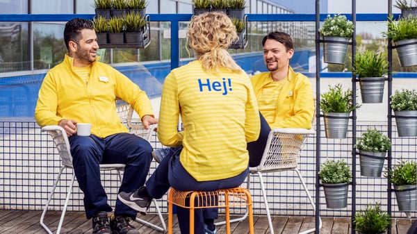 Tri osobe sede na drvenoj terasi na otvorenom, nose žute jakne, a osoba u sredini ima natpis 