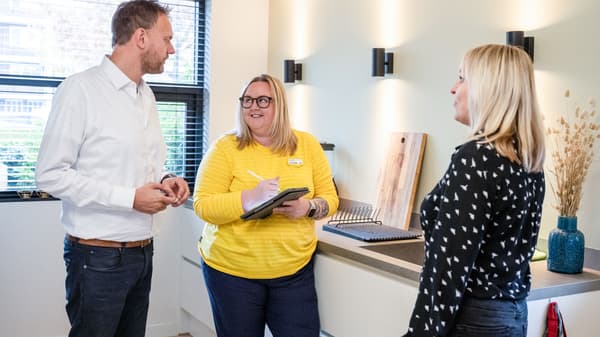 Tri osobe stoje u modernoj kuhinji i razgovaraju. Jedna osoba drži bilježnicu i piše, dok su na radnoj ploči uz zid drvene daske i uz prozor vaza s ukrasnim granama. Na zidu su crne zidne lampe.