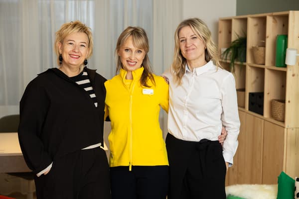 Trīs sievietes smaida kamerā, stāvot no jauna iekārtotā telpā. Vidū IKEA darbiniece dzeltenā jakā.