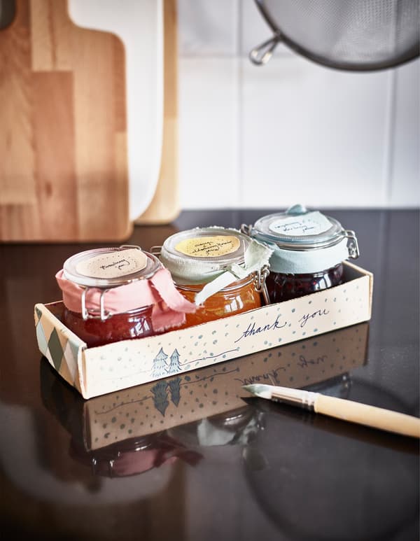 Trois bocaux de confiture décorés de rubans et placés dans un ravier en carton sur lequel il est écrit 