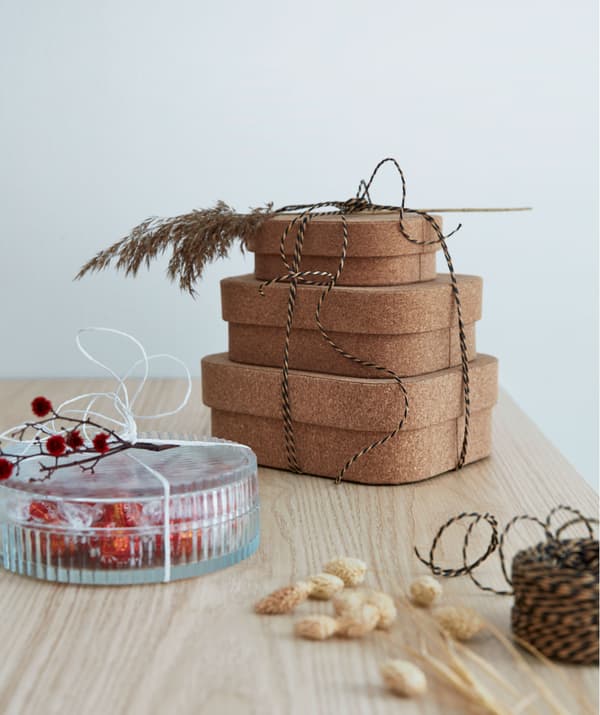 Trois boîtes en liège superposées et attachées par une ficelle, décorées avec une tige d’herbe séchée, à côté d’une boîte d’exposition en verre contenant des bonbons présentée comme un cadeau.