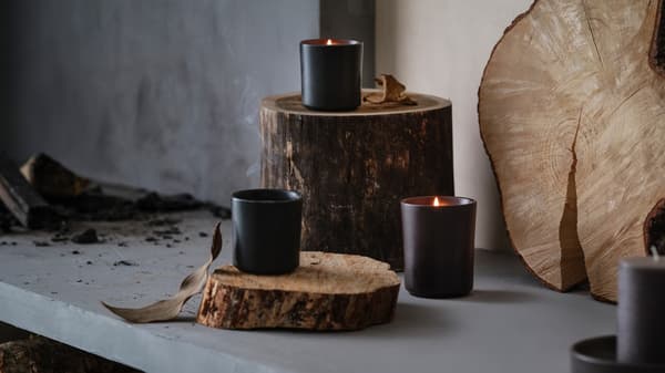 Trois bougies parfumées BEHJÄRTAD dans des pots en céramique se trouvent sur des bûches au-dessus d'un foyer en pierre rustique qui contient du bois.
