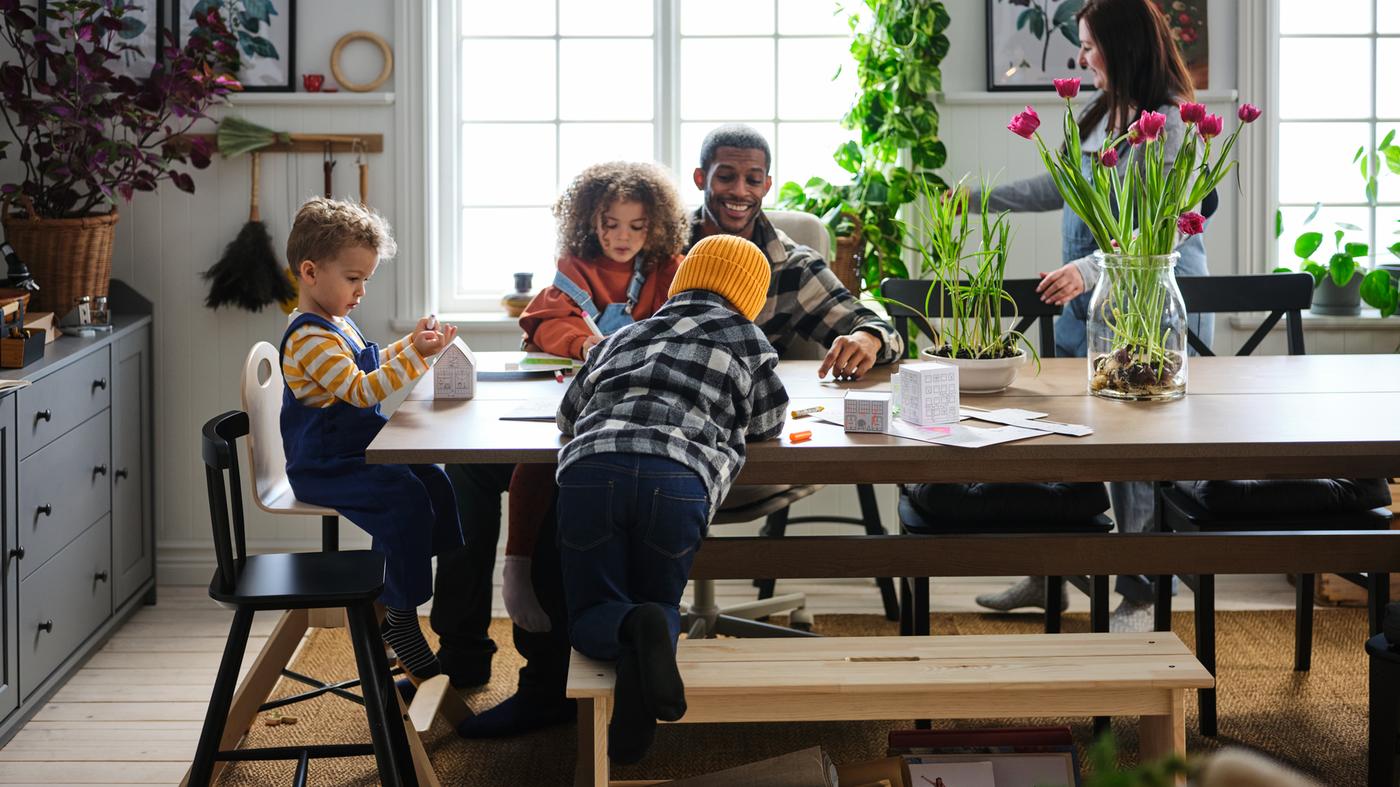 Trois jeunes enfants et leurs parents vaquent gaiement à leurs occupations dans une salle à manger avec table KLIMPFJÄLL gris-brun. 