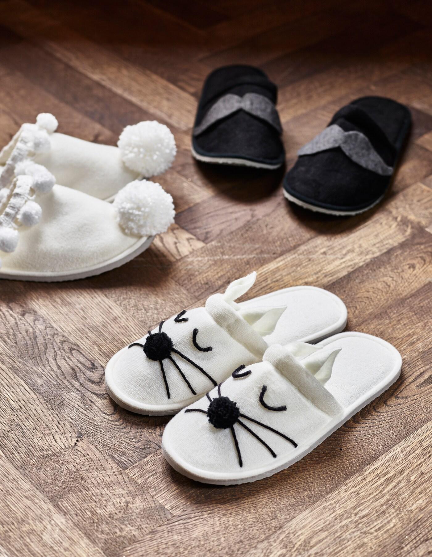 Trois paires de pantoufles TASJON customisées et posées sur un sol en parquet marron : une paire blanche avec des petits ponpons, une paire blanche avec des moustaches et yeux fait en fil de laine et une paire noire avec des petites moustaches en laine 