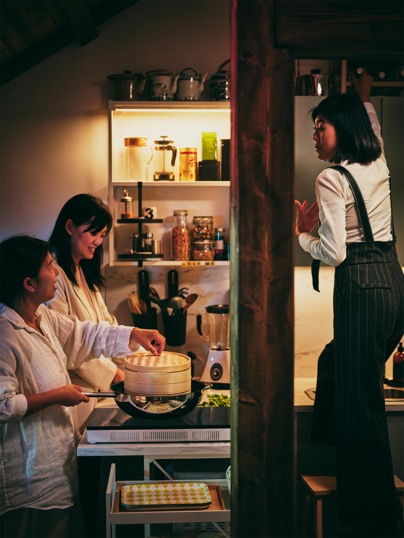Trois personnes dans une cuisine, dont une qui fait cuire de la nourriture à la vapeur au-dessus d’un wok IKEA 365+ sur une table de cuisson à induction portative TILLREDA.