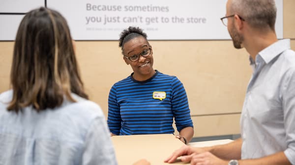 Troje ljudi razgovara oko stola u trgovini; osoba u sredini nosi plavu prugastu majicu i bedž.