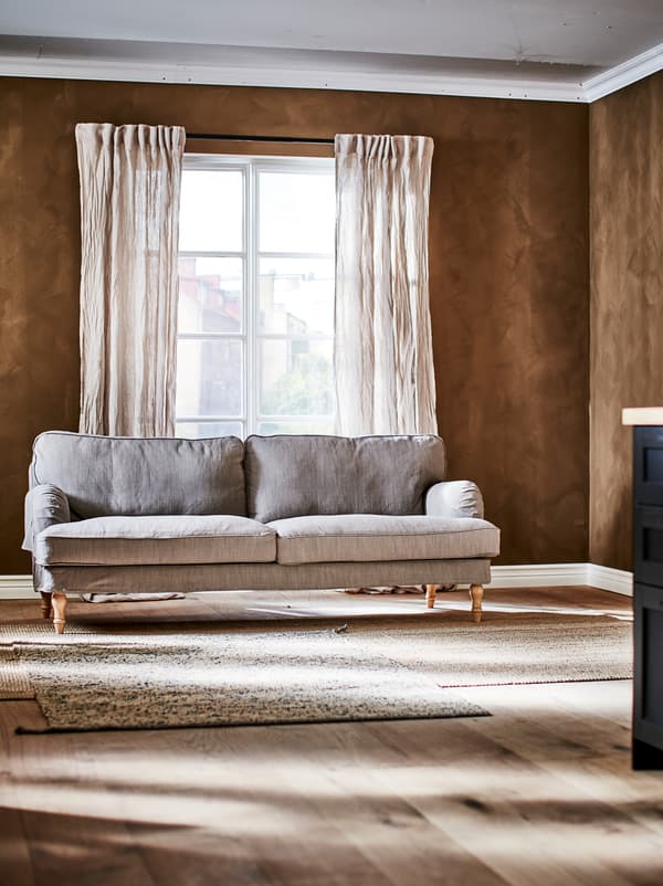Trojmiestna pohovka v hnedej izbe s francúzskymi oknami, pokrčenými ľanovými závesmi a kobercom MELHOLT na drevenej podlahe.