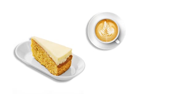 Trojúhelníček mrkvového dortu s bílou polevou na bílém oválném talířku a cappuccino v hrníčku s podšálkem