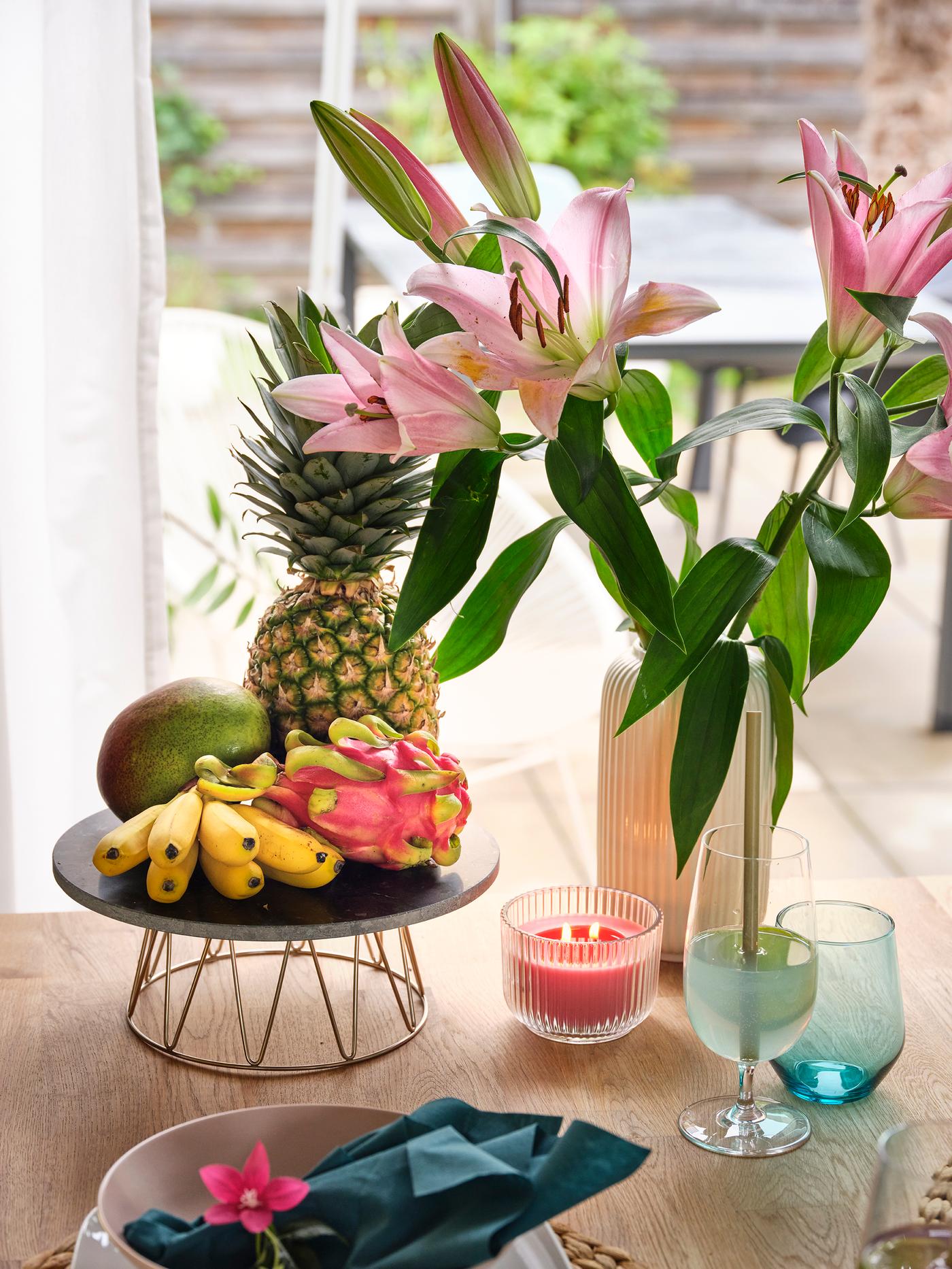Tropical table decor with pink lilies, pineapple, dragon fruit, bananas, candle and colourful glasses.