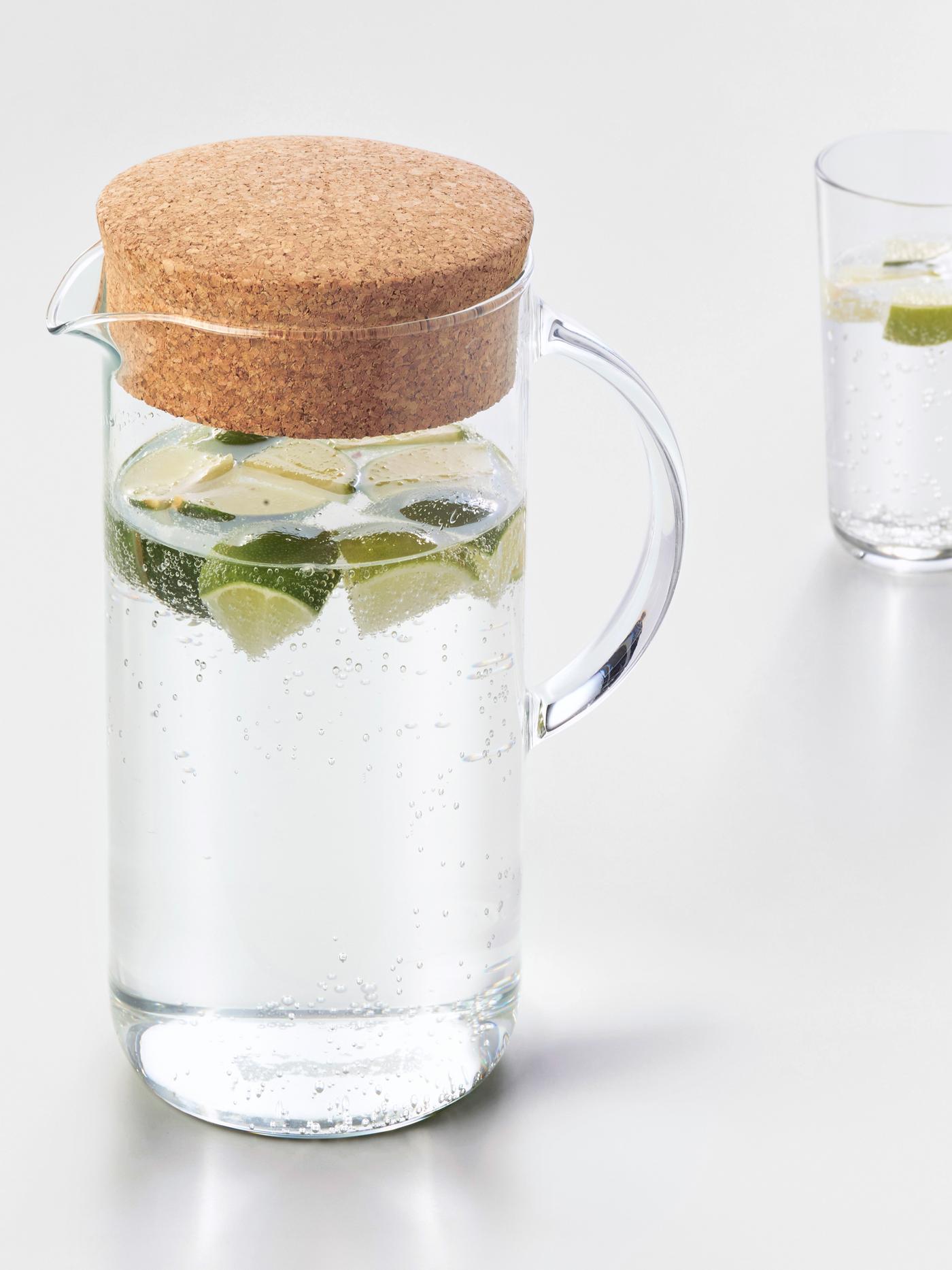 Trozos de limón flotan en el agua dentro de una jarra IKEA 365+ de vidrio con tapa de corcho y un vaso alto detrás, ambos sobre una superficie blanca.