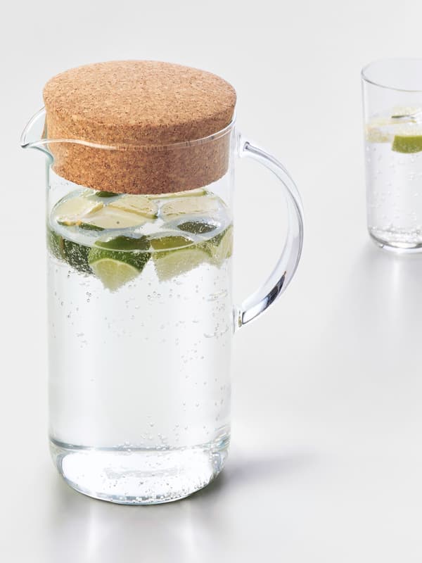 Trozos de limón flotan en el agua dentro de una jarra IKEA 365+ de vidrio con tapa de corcho y un vaso alto detrás, ambos sobre una superficie blanca.