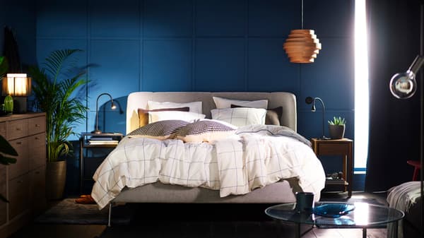 TUJFORD upholstered bed frame with BREDVECKLARE duvet cover layered on top of it, within a bedroom with dark blue walls. A foldable bedside table sits on top of the bed with a black and white chess set. On the left hand corner, a DYKARKLOCKA pendant lamp shade in white stained oak veneer hangs from the ceiling. Black out MAJGULL curtains are pulled apart slightly to let some light in.