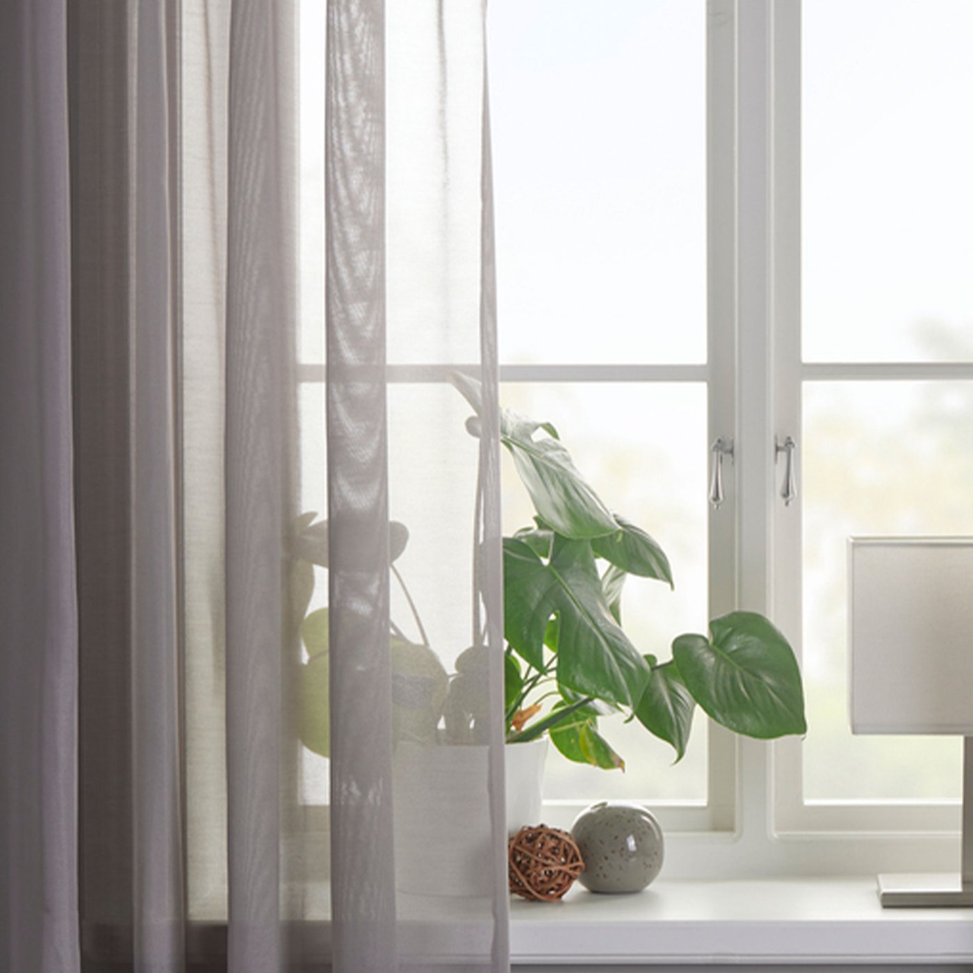 Tunn grå gardin i fönster med grön växt och vit lampa.