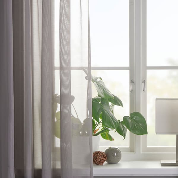 Tunn grå gardin i fönster med grön växt och vit lampa. 