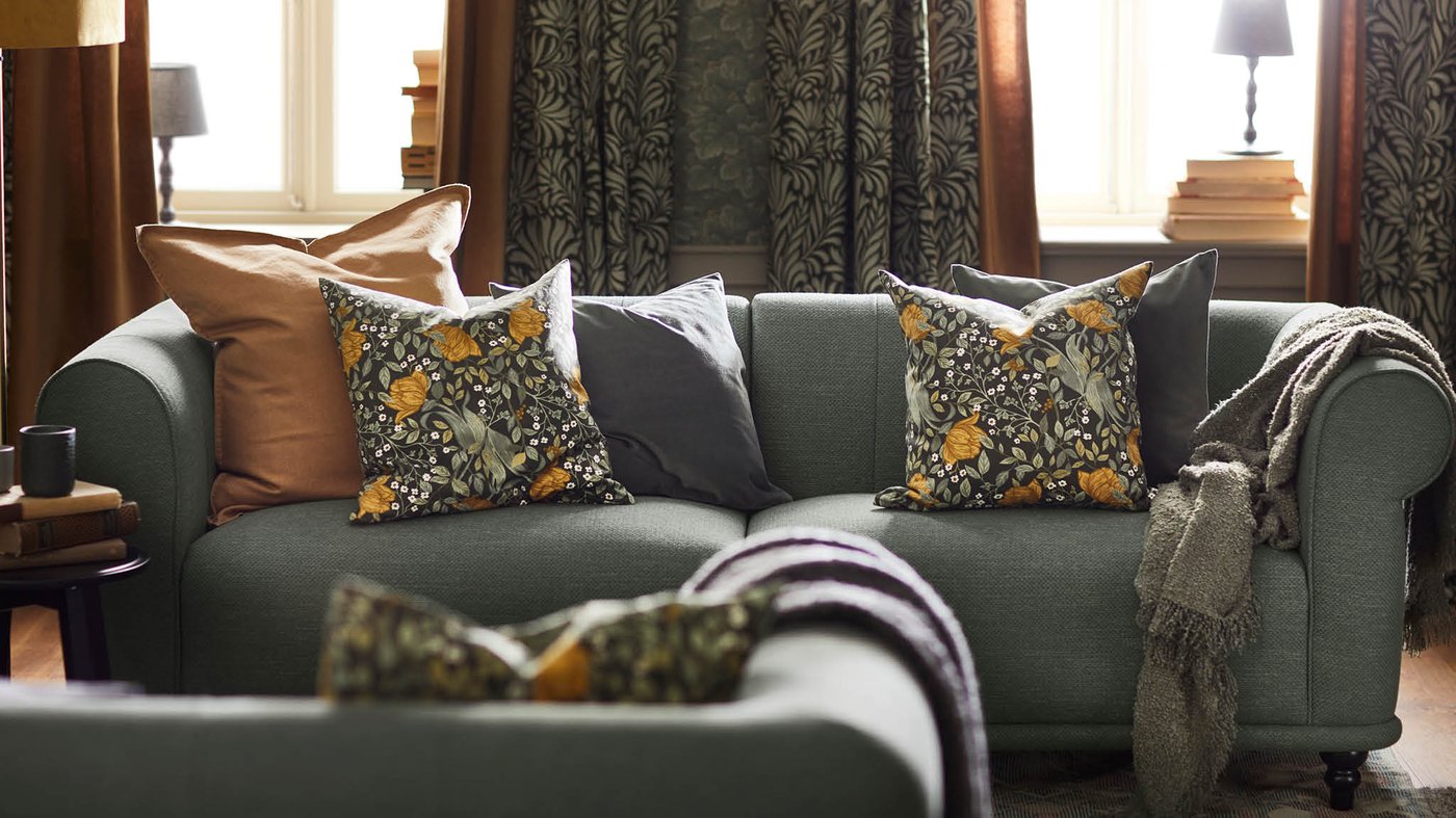 TUVSÄV díszpárnák és egy zöld SANDBRODD takaró hever egy zöld VISKAFORS kanapén. A háttérben ablakok, az ablakok előtt pedig TRÅGSPINNARE függönyök.