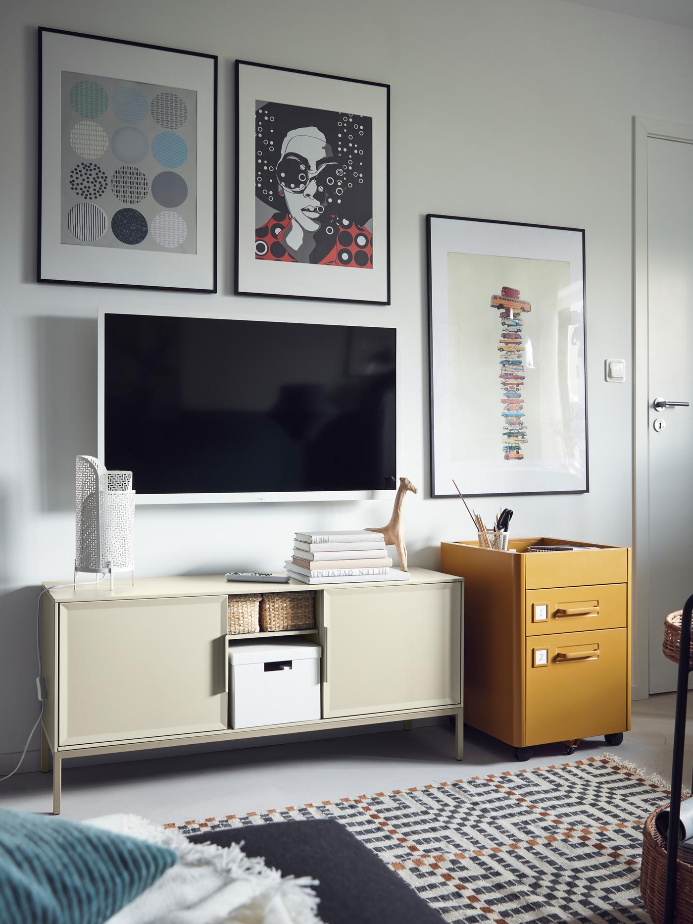 TV et images sur un mur au-dessus d’une armoire métallique TULLSTORP beige avec rangement ouvert accueillant des paniers et une boîte.