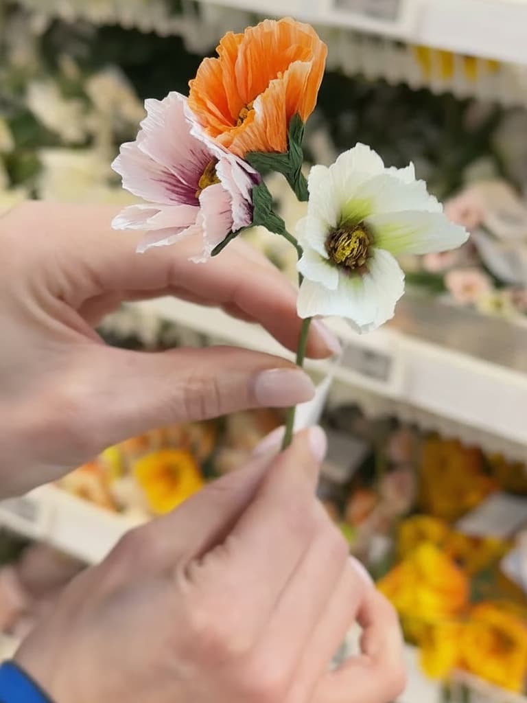 Två händer håller upp en bukett med tre pappersblommor – en orange, en ljusrosa och en vit – framför en suddig bakgrund av andra blommor i en butik.