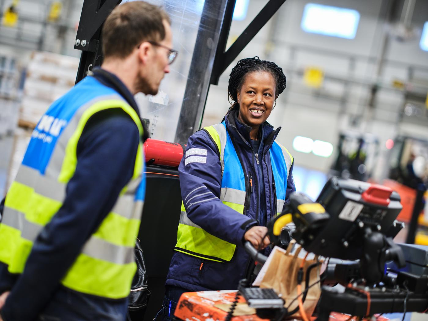 Två IKEA medarbetare med säkerhetsvästar på sig är i ett lager och står vid en truck.