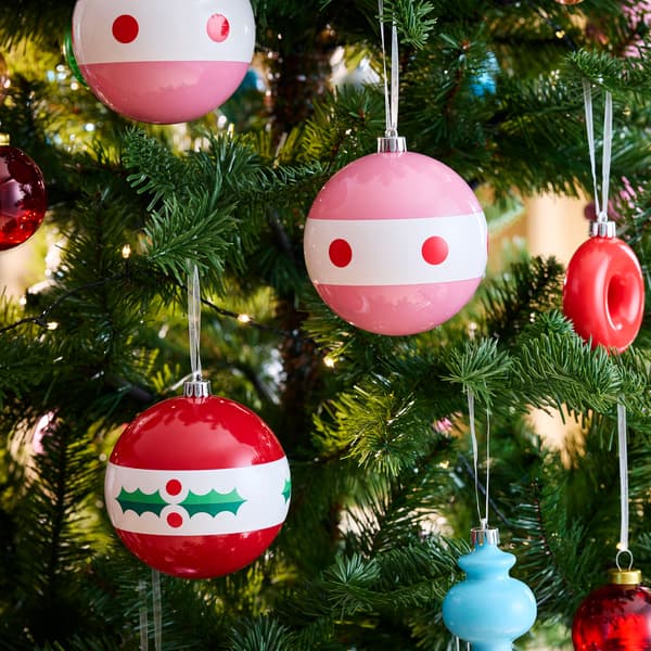 Två julgransdekorationer hängande från grenar; en röd med vita och gröna detaljer, och en rosa med vita inslag.