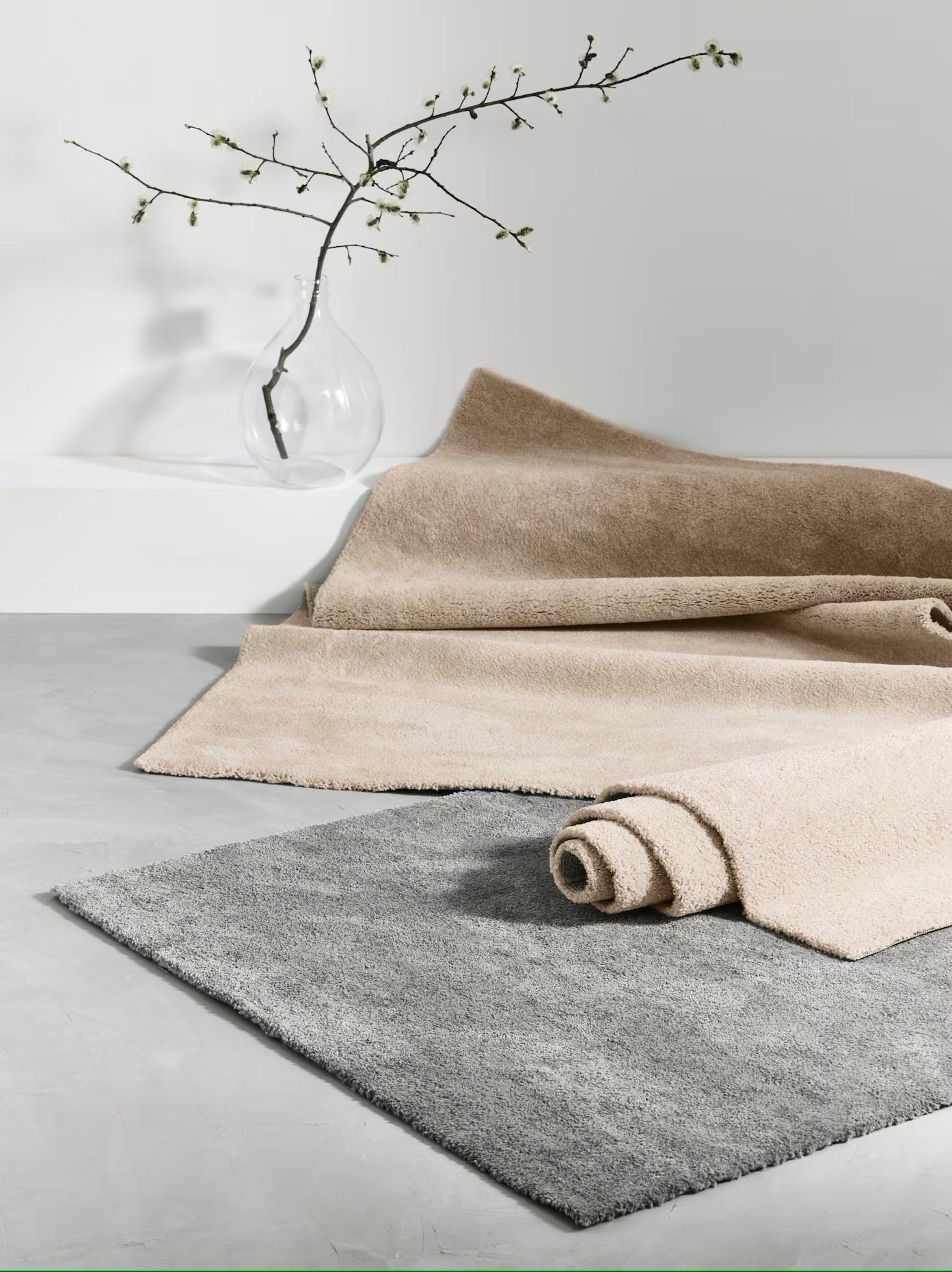 Två mattor, en beige delvis upprullad och en grå, på ljust golv med glasvas och gren i bakgrunden.