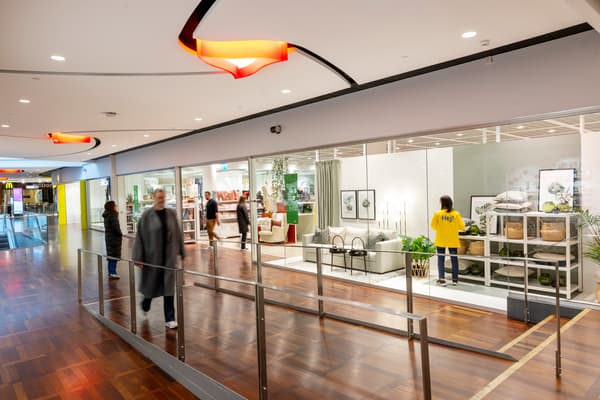 Två mörkklädda människor går förbi i en galleria framför ett skyltfönster. Där inne syns medarbetare i gula kläder