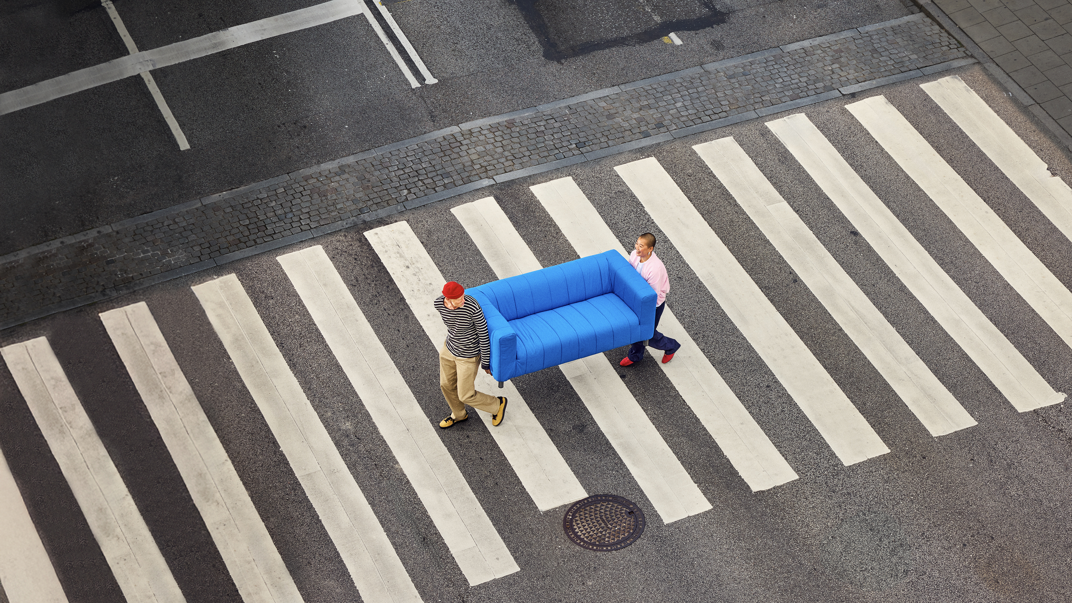 Två personer går på ett övergångsställe. De bär på en blå soffa. 
