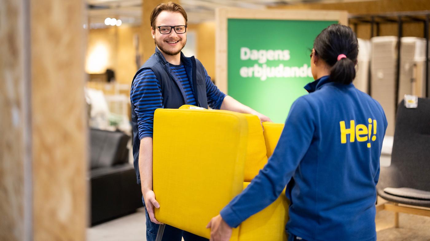 Två personer i blåa IKEA-kläder bär en gul fåtölj. 