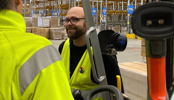 Två personer i varseljackor i ett lager, med hyllor, lådor och utrustning i bakgrunden.