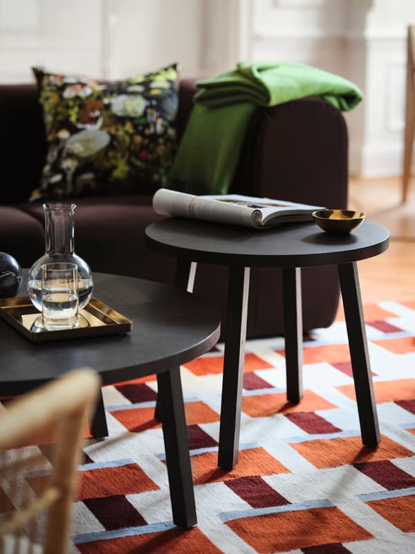 Två svarta runda bord i ett vardagsrum, med en tidning och en liten skål ovanpå. En glasvas står på ett bricka på det andra bordet. En färgglad kudde och en grön filt ligger på en brun soffa i bakgrunden.