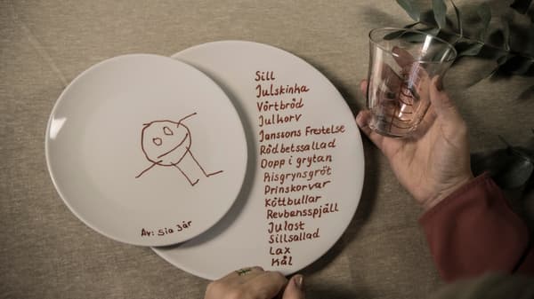 Två tallrikar på en duk och en hand som håller i ett glas. Alla tre har det målats och skrivits på med porslinspenna. 
