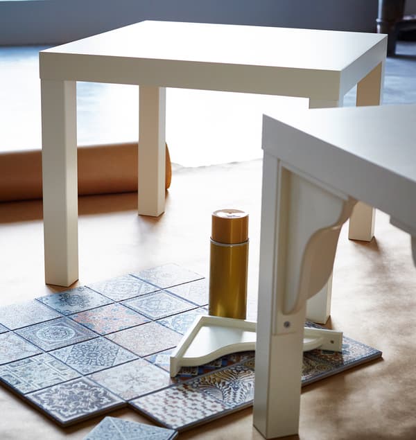 Två vita IKEA LACK bord redo att hackas, medan kakelplattorna arrangeras på golvet intill dem.
