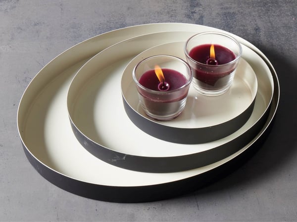 Twee brandende paarse kaarsen in glazen houders staan samen op een set stapelbare dienbladen die doen denken aan een minimalistische IKEA‑presentatie.