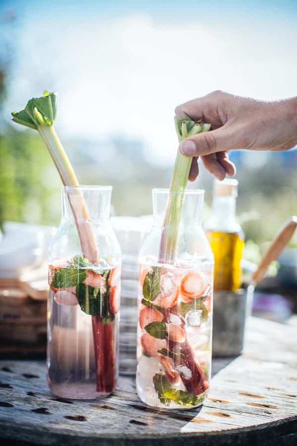 Twee glazen flessen gevuld met een drank met aardbeien, muntblaadjes en rabarberstelen staan op een tafel, terwijl een hand een van de rabarberstelen in de rechterfles vasthoudt.

