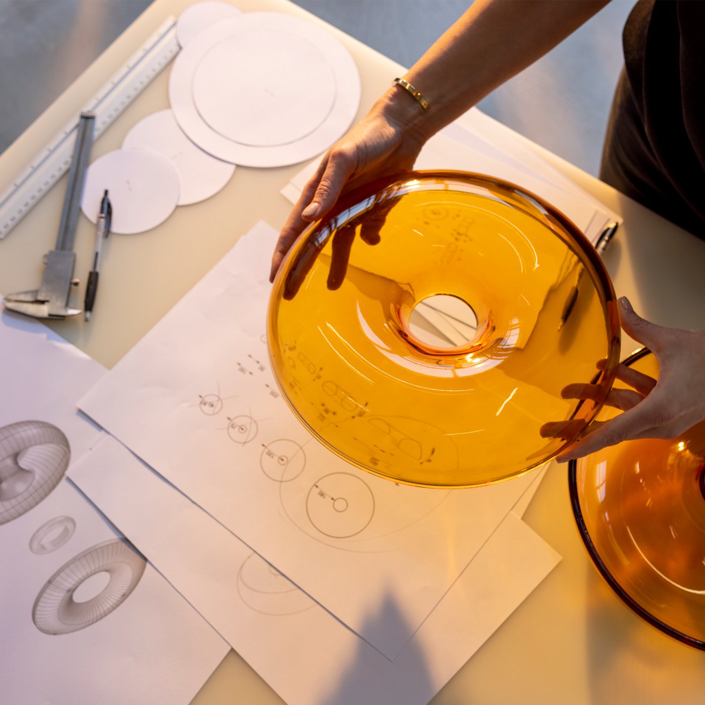 Twee handen houden het deksel van een oranje, glazen VARMBLIXT donutvormige schaal omhoog boven een tafel met schetsen van het object.