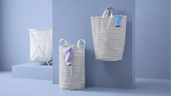 Twee KLUNKA waszakken en een opvouwbare wasmand voor een blauwe muur. De ene waszak staat op zichzelf, de andere wastas hangt aan een haakje aan de muur. Multi-inzetbaar en functioneel.