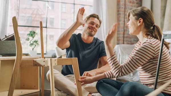 Twee mensen die samen op de vloer meubels monteren en elkaar een high‑five geven in een lichte kamer met bakstenen muur.