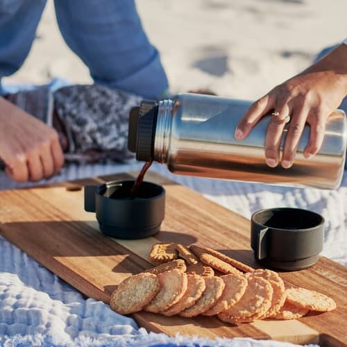 Twee mensen op een picknickkleed terwijl een van hen koffie uit een stalen isoleerfles schenkt in zwarte herbruikbare bekers.