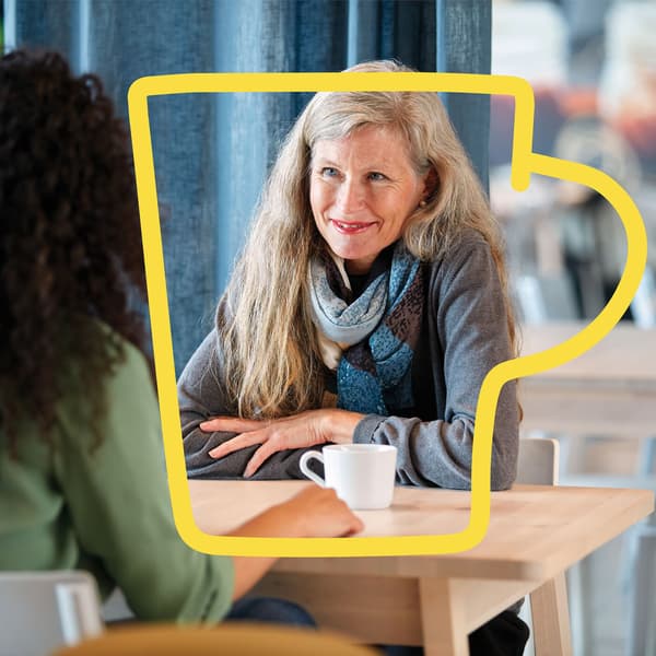 Twee mensen zitten aan tafel in café; één met grijze trui en sjaal houdt witte koffiekop vast, gezicht is wazig.