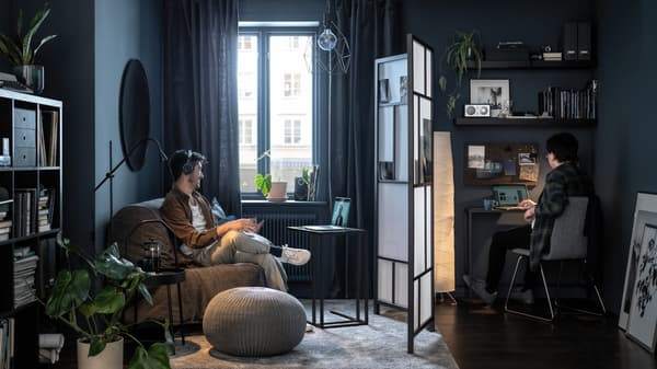 Twee mensen zitten aan weerszijden van een woonkamer, gescheiden door een RISÖR scheidingswand, en kijken allebei naar hun scherm.