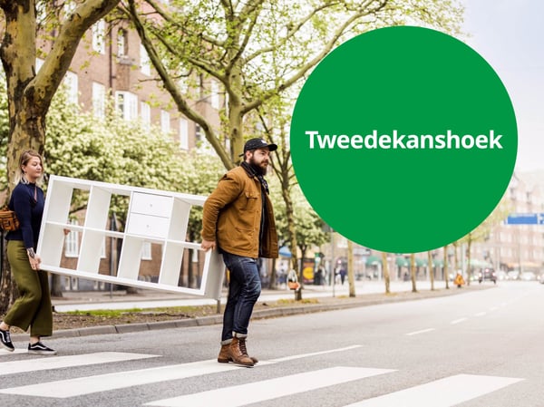 Twee personen dragen samen een wit opbergmeubel over een zebra­pad in een stadsstraat met bomen, naast een groen rond label met de tekst “Tweedekanshoek”.
