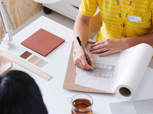 Twee personen zitten aan een tafel. De ene tekent een kaart op papier en de andere kijkt toe. Op de tafel staan onder meer twee kopjes thee, een laptop, kleurstalen, een liniaal en diverse documenten.
