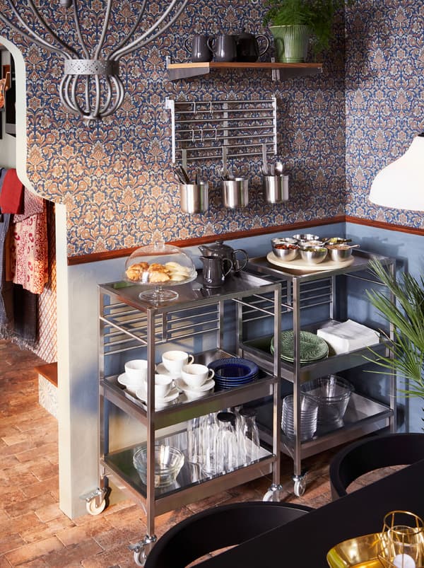 Twee roestvrij stalen KUNGSFORS roltafels vormen een onbijtbuffet met glazen, een opdienschaal en ander servies. Daarboven hangen potten met bestek.
