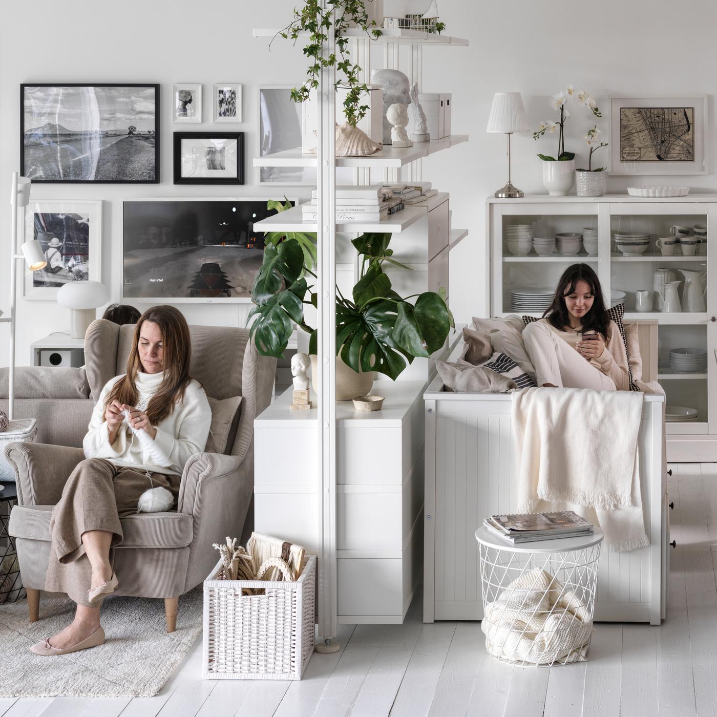 Twee vrouwen zitten aan weerskanten van een ELVARLI opbergelementen in een ruime, lichte woonkamer. Een van de vrouwen zit in een beige STRANDMON fauteuil.