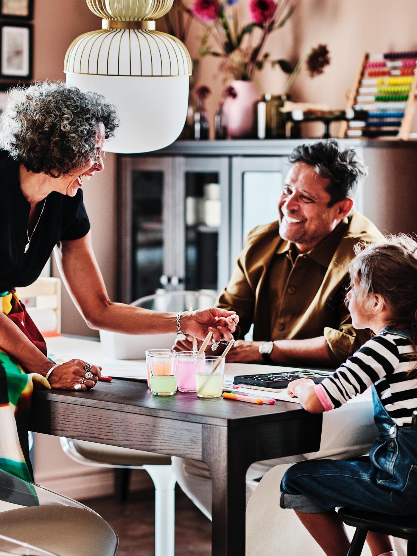 Two adults and a child around a dark brown EKEDALEN extendable table. The child sits on a black AGAM junior chair.
